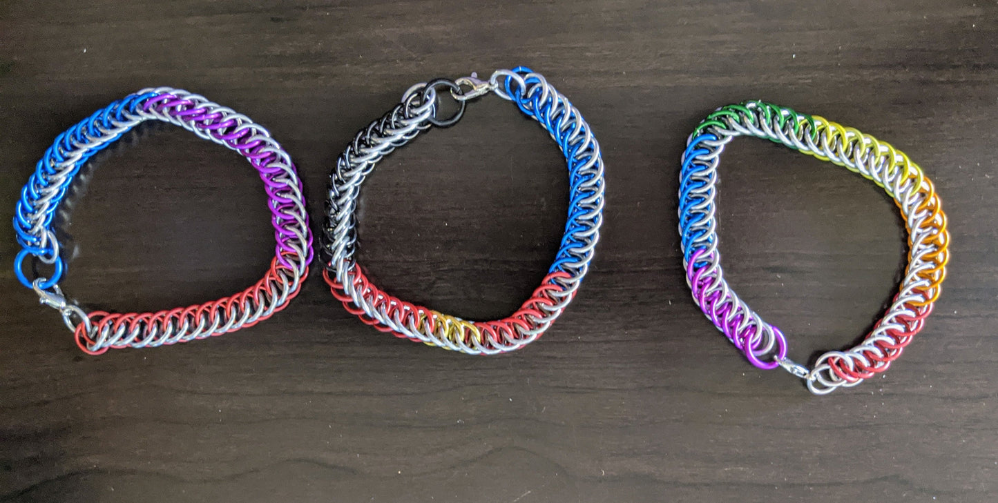 Three chainmail bracelets displayed side by side on a wooden surface. Each bracelet has a different color pattern; from left to right, blue and purple, green and orange, and red and yellow.