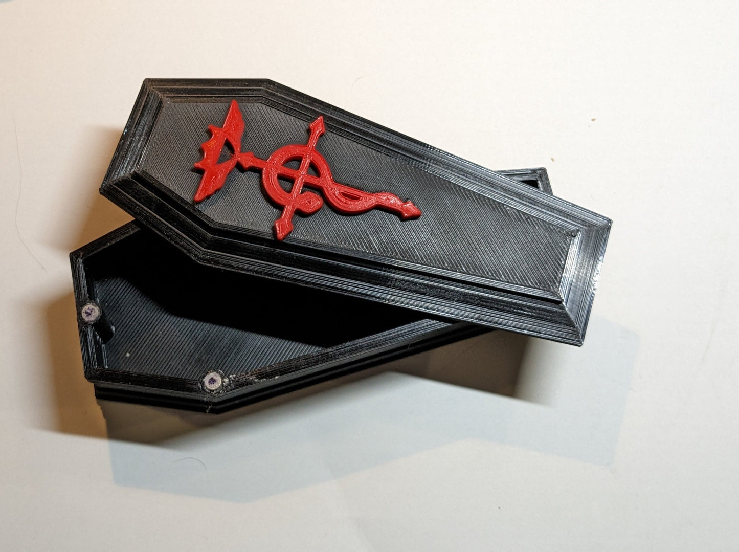 3D printed coffin shaped dice/trinket box in black with a red alchemy symbol in the top. The top is slightly askew to show the inside of the box