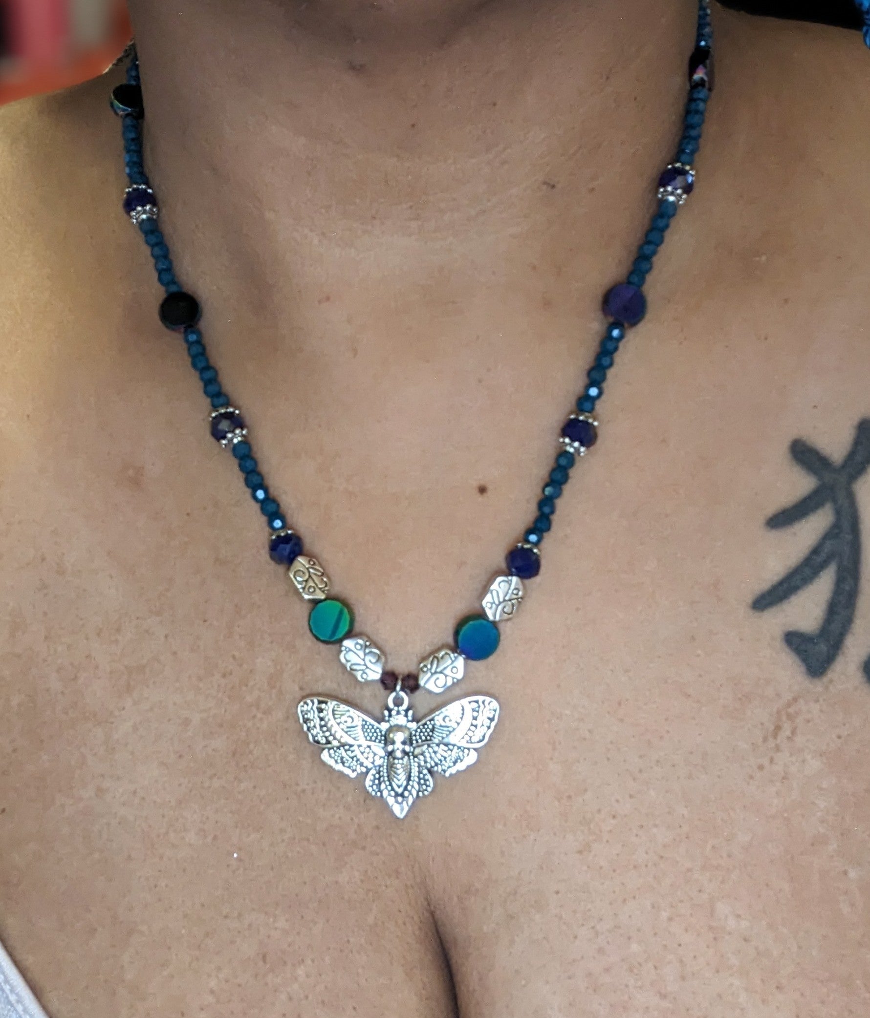 Beaded necklace with antiqued-silver colored deaths-head moth pendant.  The necklace has glass beads in shades of blue & teal with silver bead caps and engraved silver accent beads.  It has an ornate round silver magnetic clasp.  The necklace is shown around the neck of a brown person.