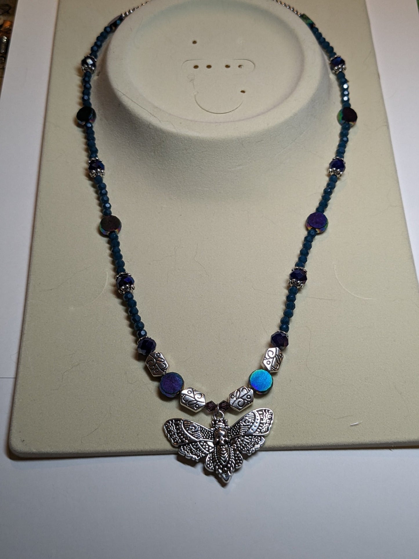 Beaded necklace with antiqued-silver colored deaths-head moth pendant. The necklace has glass beads in shades of blue & teal with silver bead caps and engraved silver accent beads. It has an ornate round silver magnetic clasp. The necklace is about 20 inches long.
