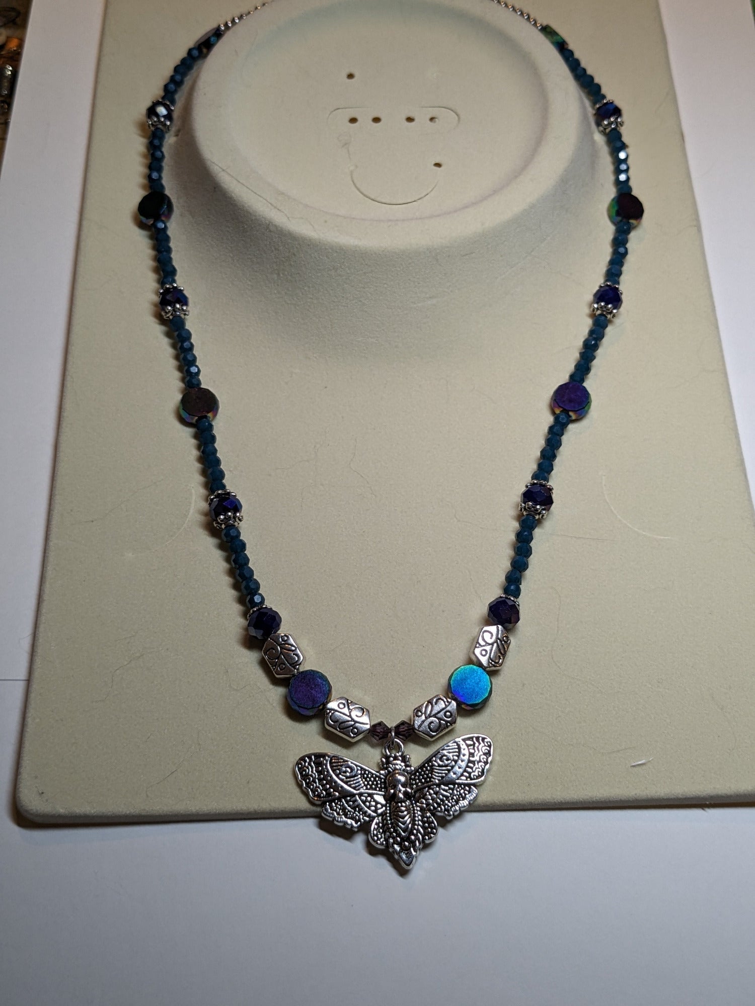 Beaded necklace with antiqued-silver colored deaths-head moth pendant.  The necklace has glass beads in shades of blue & teal with silver bead caps and engraved silver accent beads.  It has an ornate round silver magnetic clasp.  The necklace is about 20 inches long.