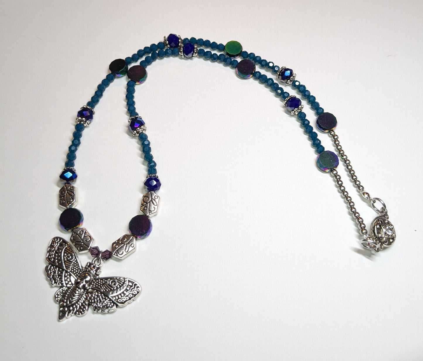 Beaded necklace with antiqued-silver colored deaths-head moth pendant. The necklace has glass beads in shades of blue & teal with silver bead caps and engraved silver accent beads. It has an ornate round silver magnetic clasp. The necklace is about 20 inches long.