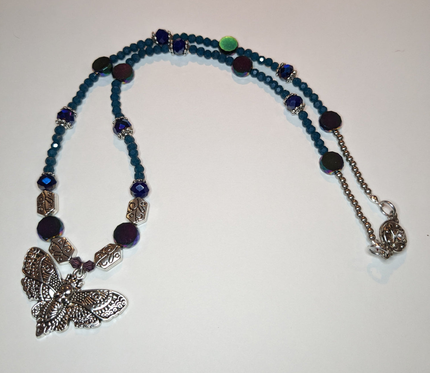 Beaded necklace with antiqued-silver colored deaths-head moth pendant. The necklace has glass beads in shades of blue & teal with silver bead caps and engraved silver accent beads. It has an ornate round silver magnetic clasp. The necklace is about 20 inches long.