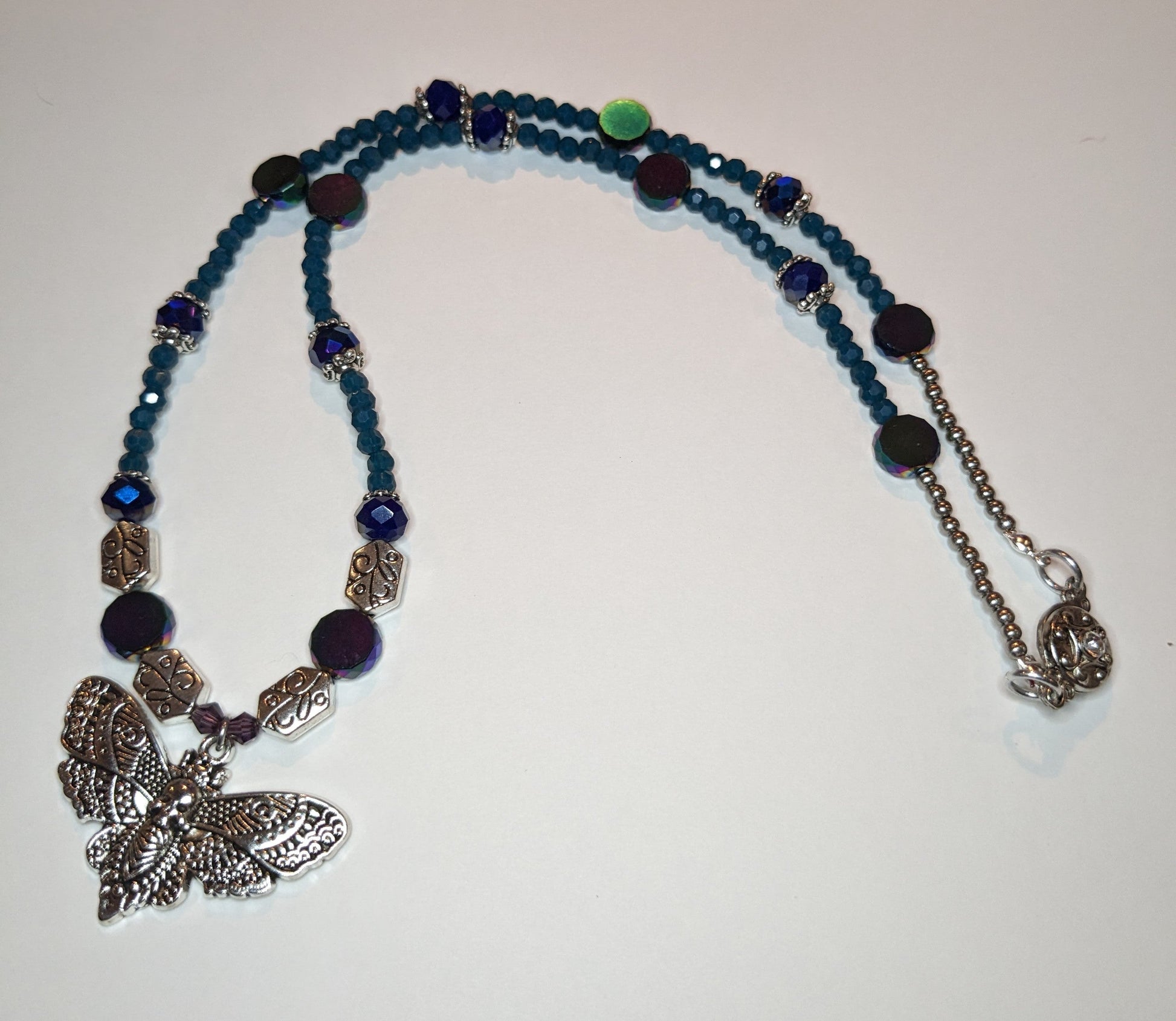 Beaded necklace with antiqued-silver colored deaths-head moth pendant.  The necklace has glass beads in shades of blue & teal with silver bead caps and engraved silver accent beads.  It has an ornate round silver magnetic clasp.  The necklace is about 20 inches long.