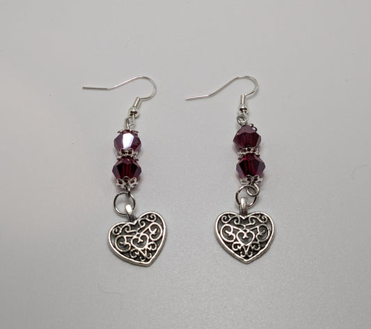 A pair of heart-shaped dangle earrings with intricate filigree design and red crystal beads on a white background.