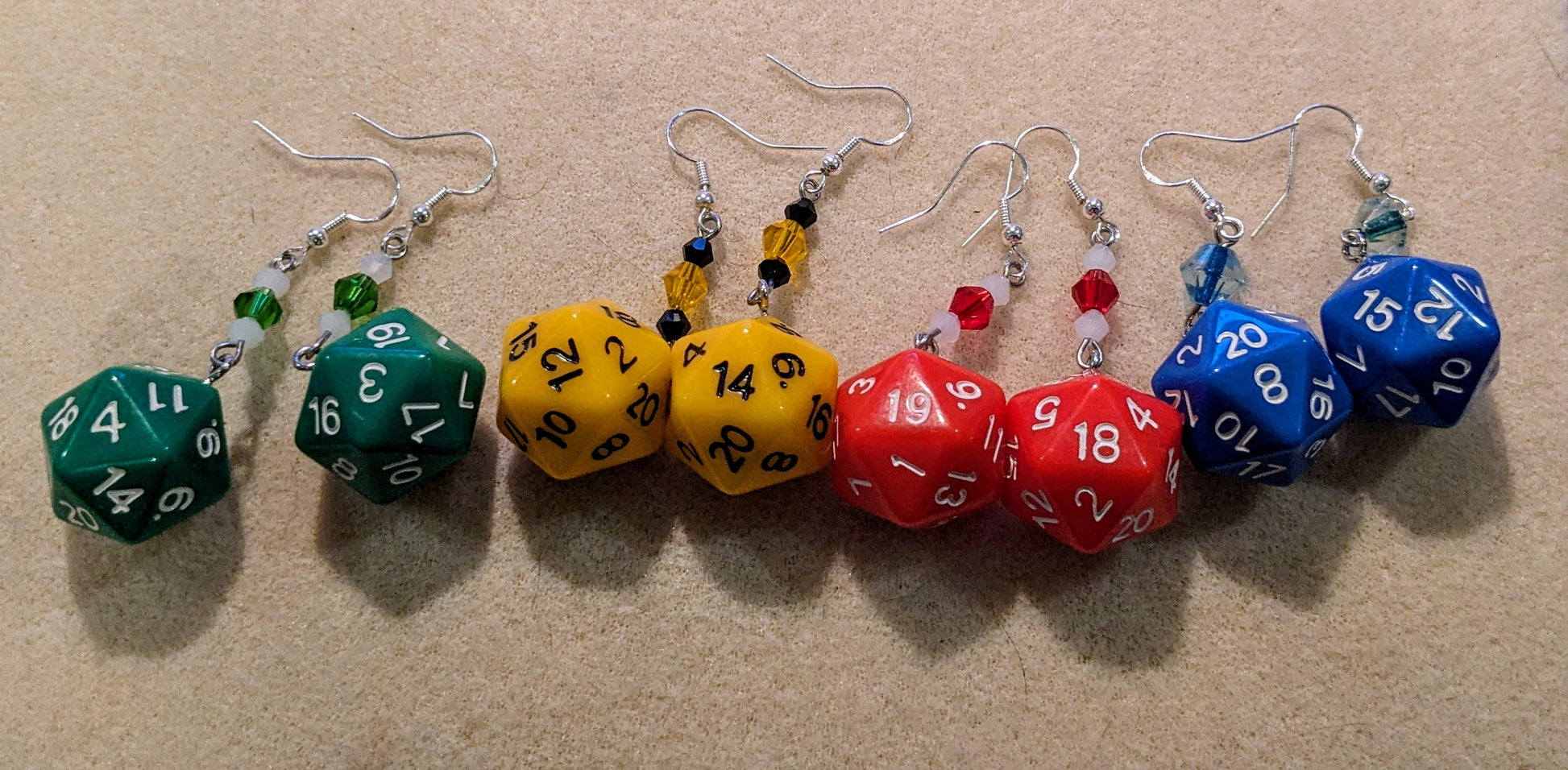 4 pairs of D20 earrings on an off-white background. Earrings are green, yellow, red & blue with wire wrapped crystals marching the dice and number colors