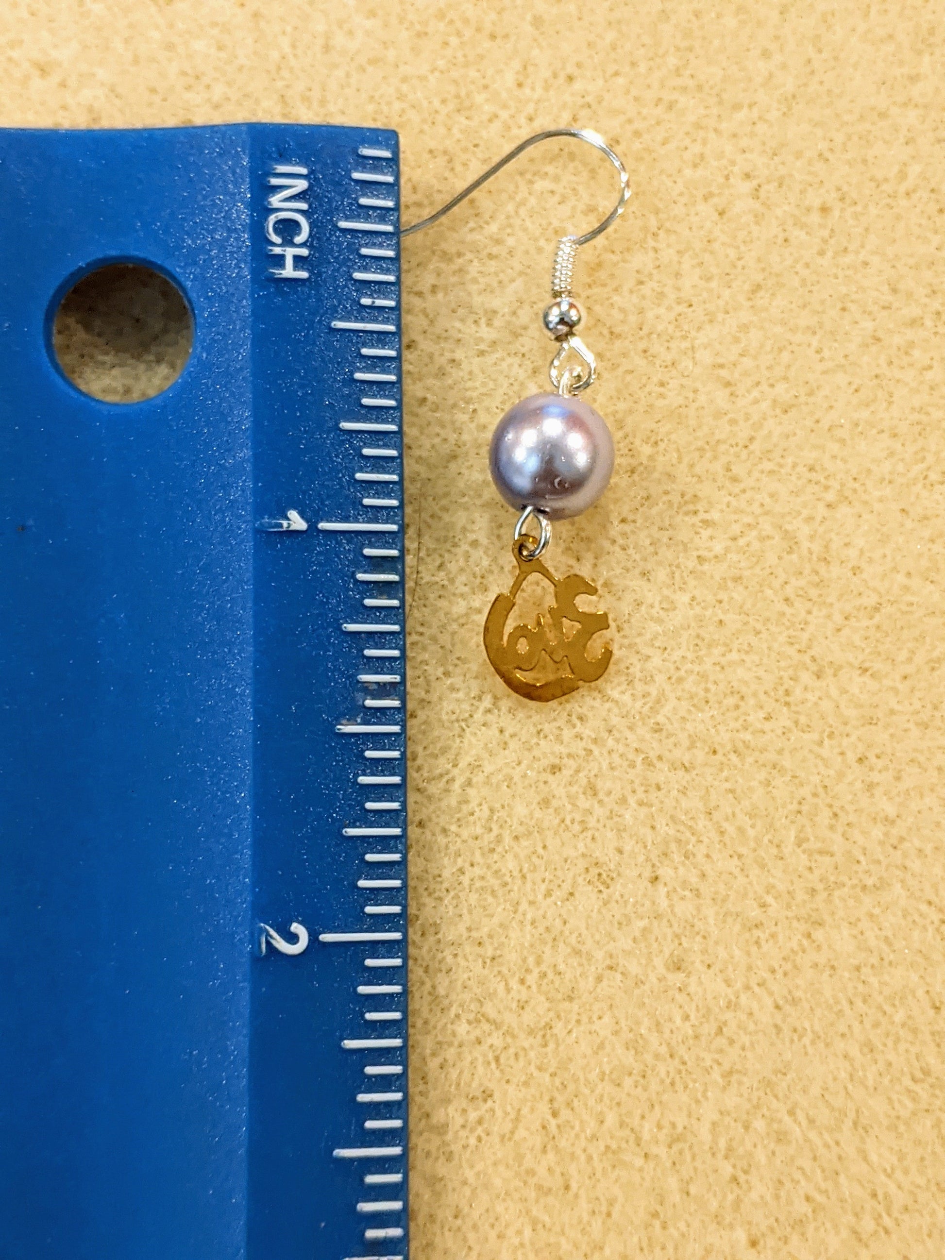 A drop earring featuring gold-colored cut-out charm that says love and pearl bead next to a blue ruler showing the earring is 1.5 inches including the ear wire