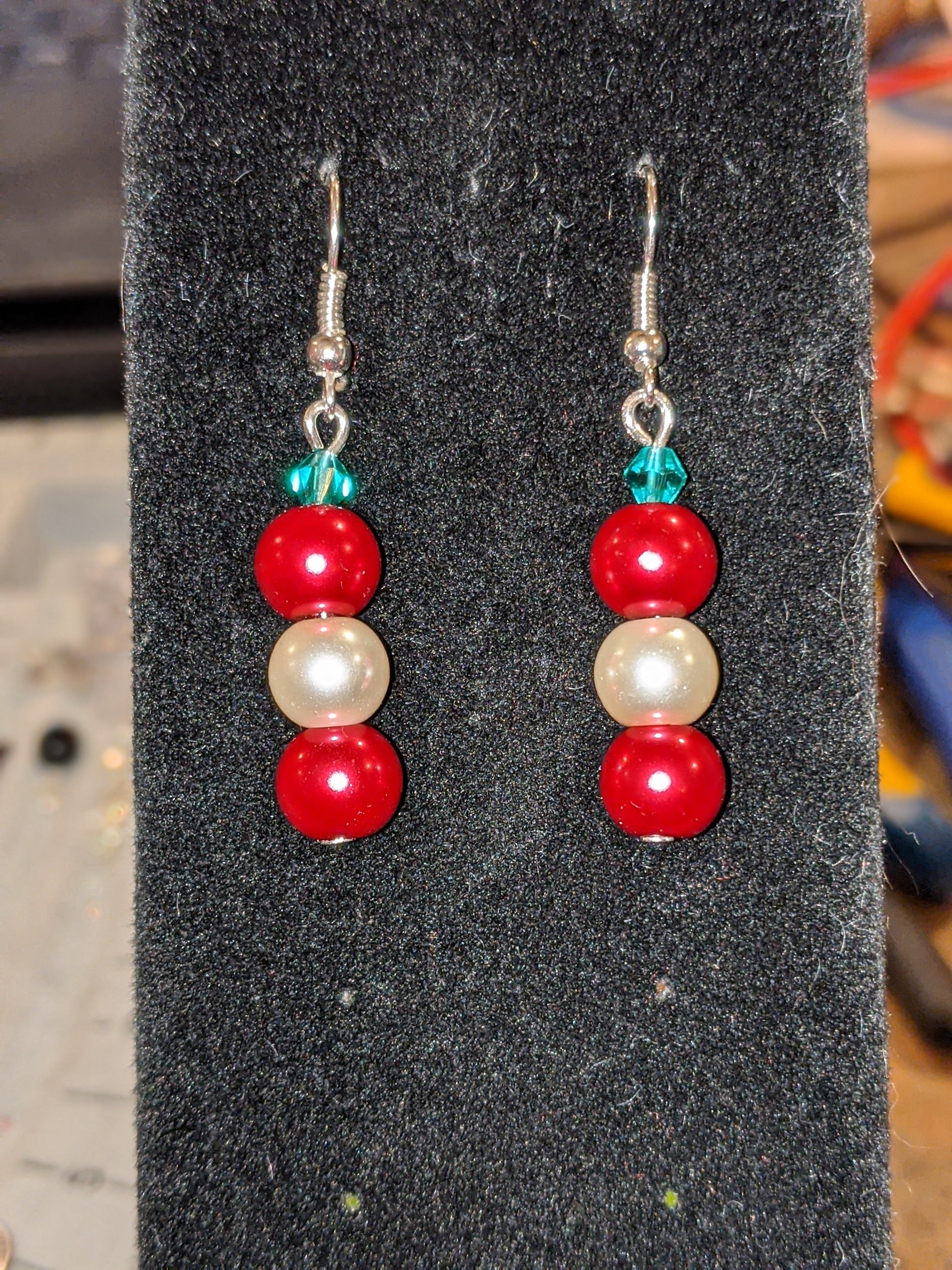 A pair of pearl bead dangle earrings with red and white glass pearls and green faceted bicone, on silver French hook ear wires displayed on a black stand.