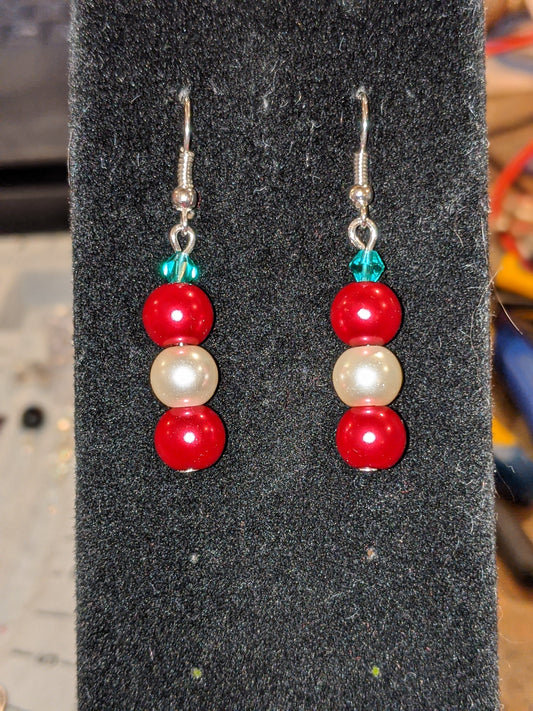 A pair of pearl bead dangle earrings with red and white glass pearls and green faceted bicone, on silver French hook ear wires displayed on a black stand.