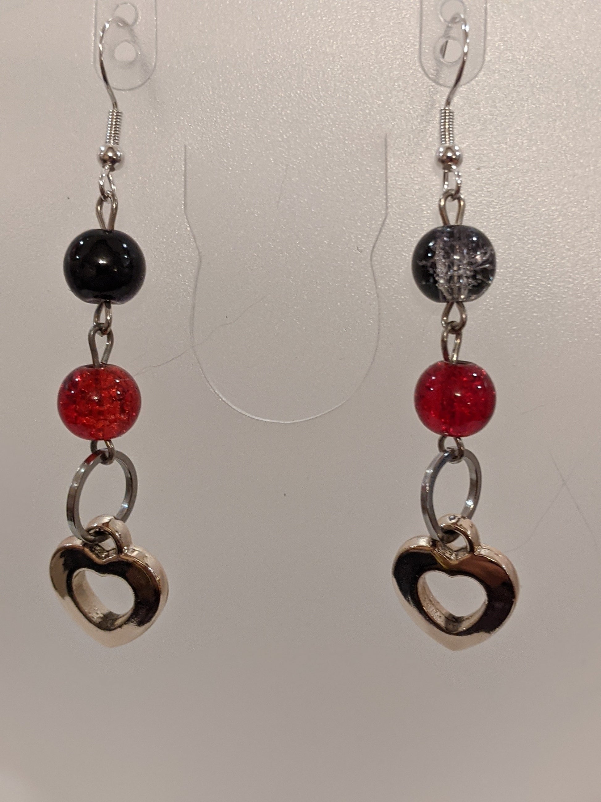 A pair of dangle earrings featuring black and red glass crackle beads with silver heart charms, dangling from silver-toned wires.