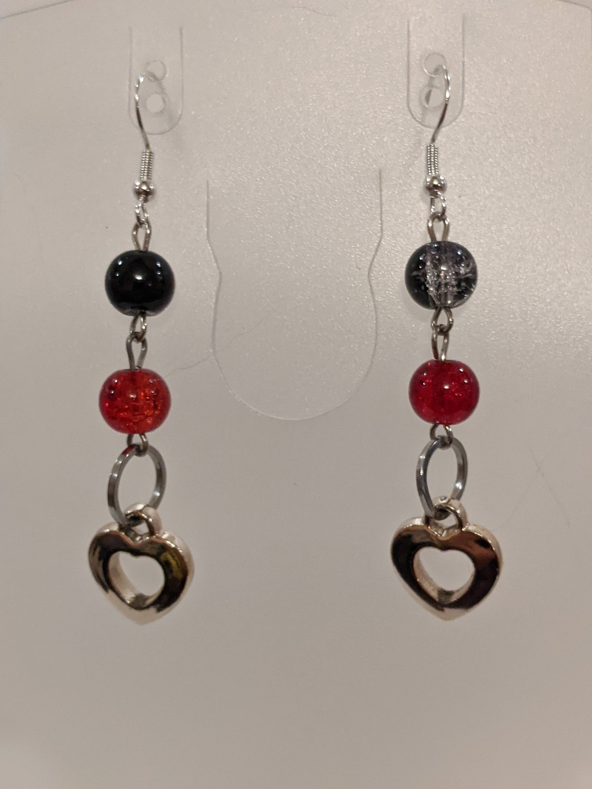 A pair of dangle earrings featuring black and red glass crackle beads with silver heart charms, dangling from silver-toned wires.
