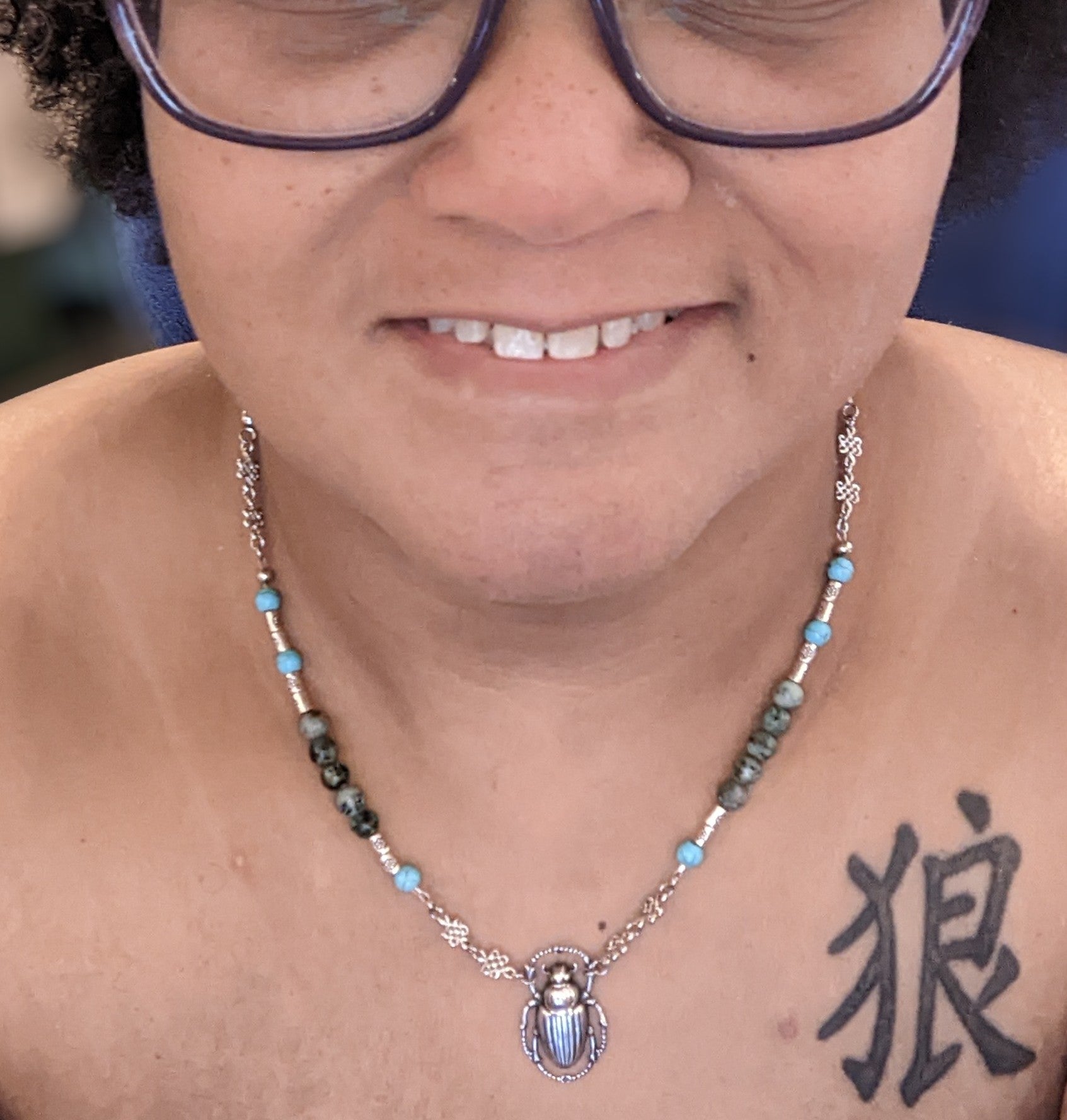 Close up of the neck of a person wearing a beaded necklace.  The necklace has a silver scarab pendant, turquoise and African turquoise beads with silver accents.