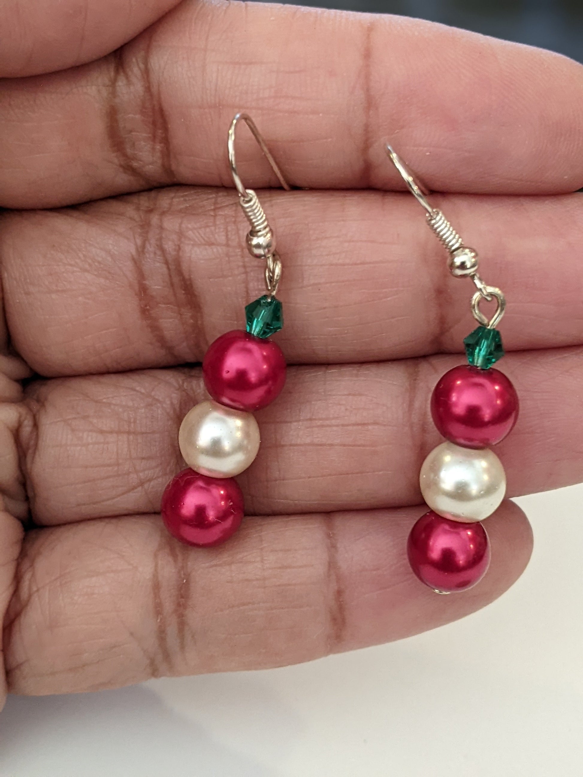 A pair of pearl bead dangle earrings with red and white glass pearls and green faceted bicone, on silver French hook ear wires being held by a brown person's fingers