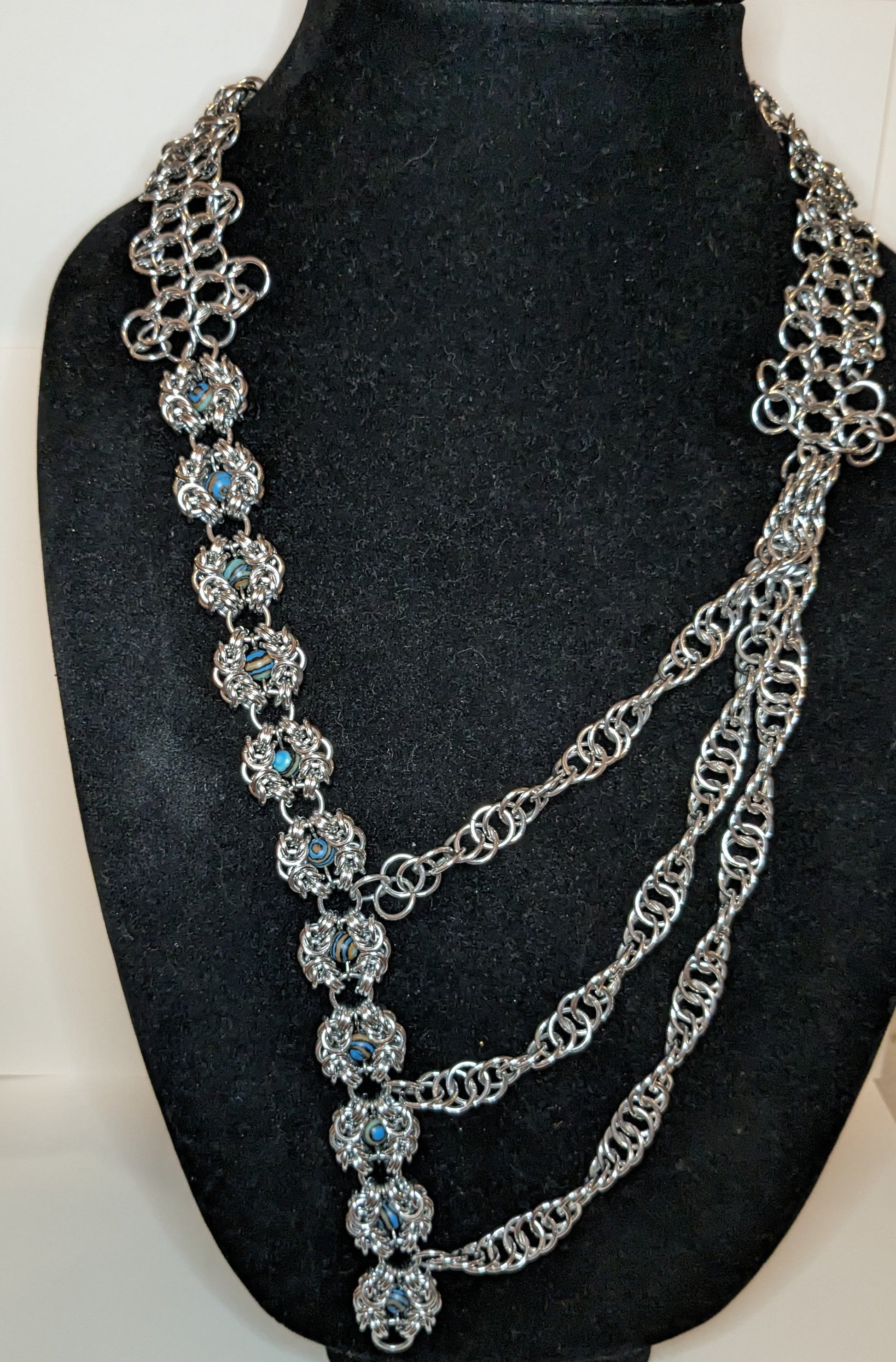 Asymmetric chainmail necklace on a black bust.  Left side is modified Romanov weave flowers with gemstone beads and the right side are three chainmail strands that cross the front to look like a mult-strand necklace.