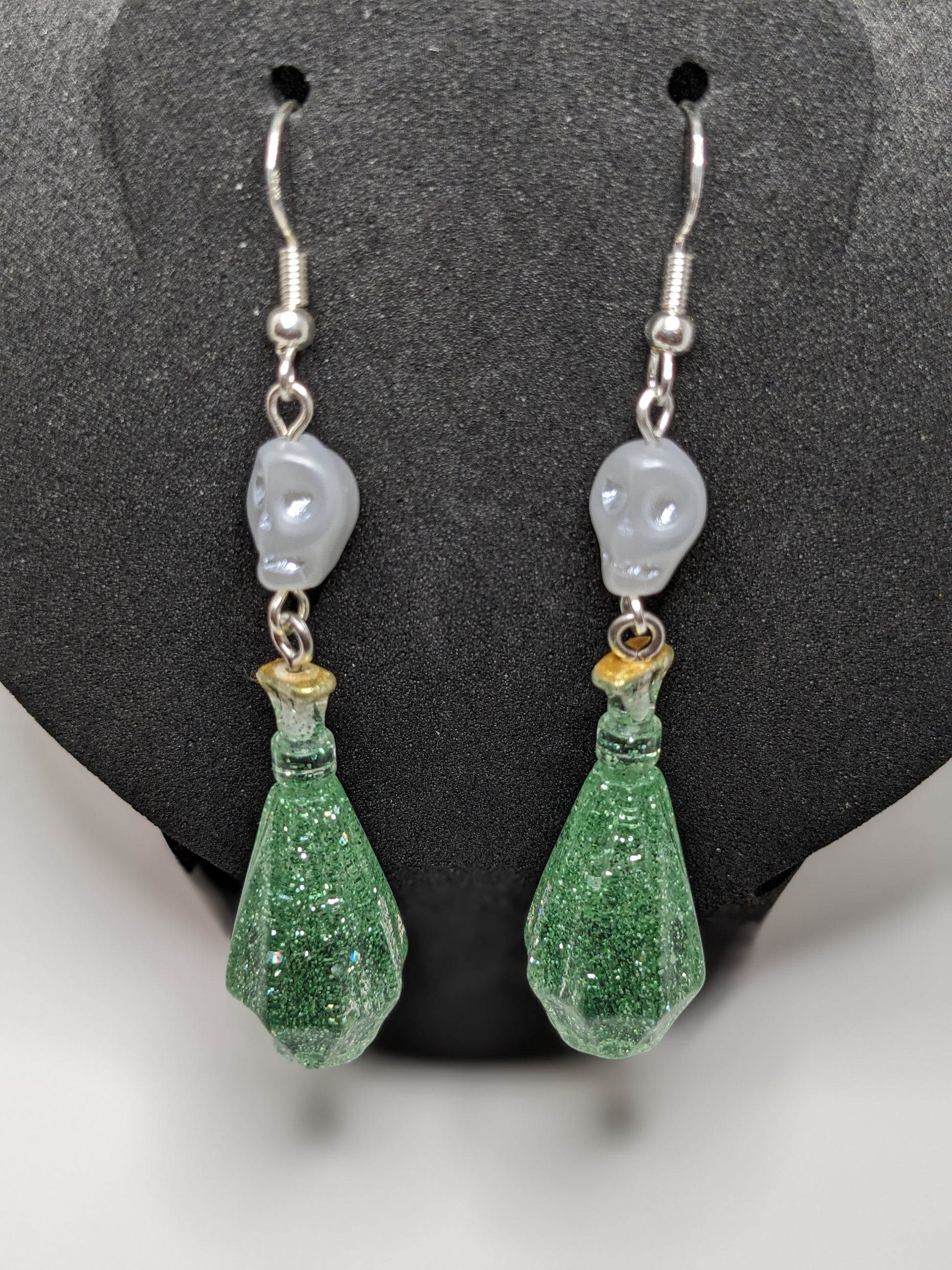 Green glittery potion bottle earrings with white skull beads on a dark gray stand.
