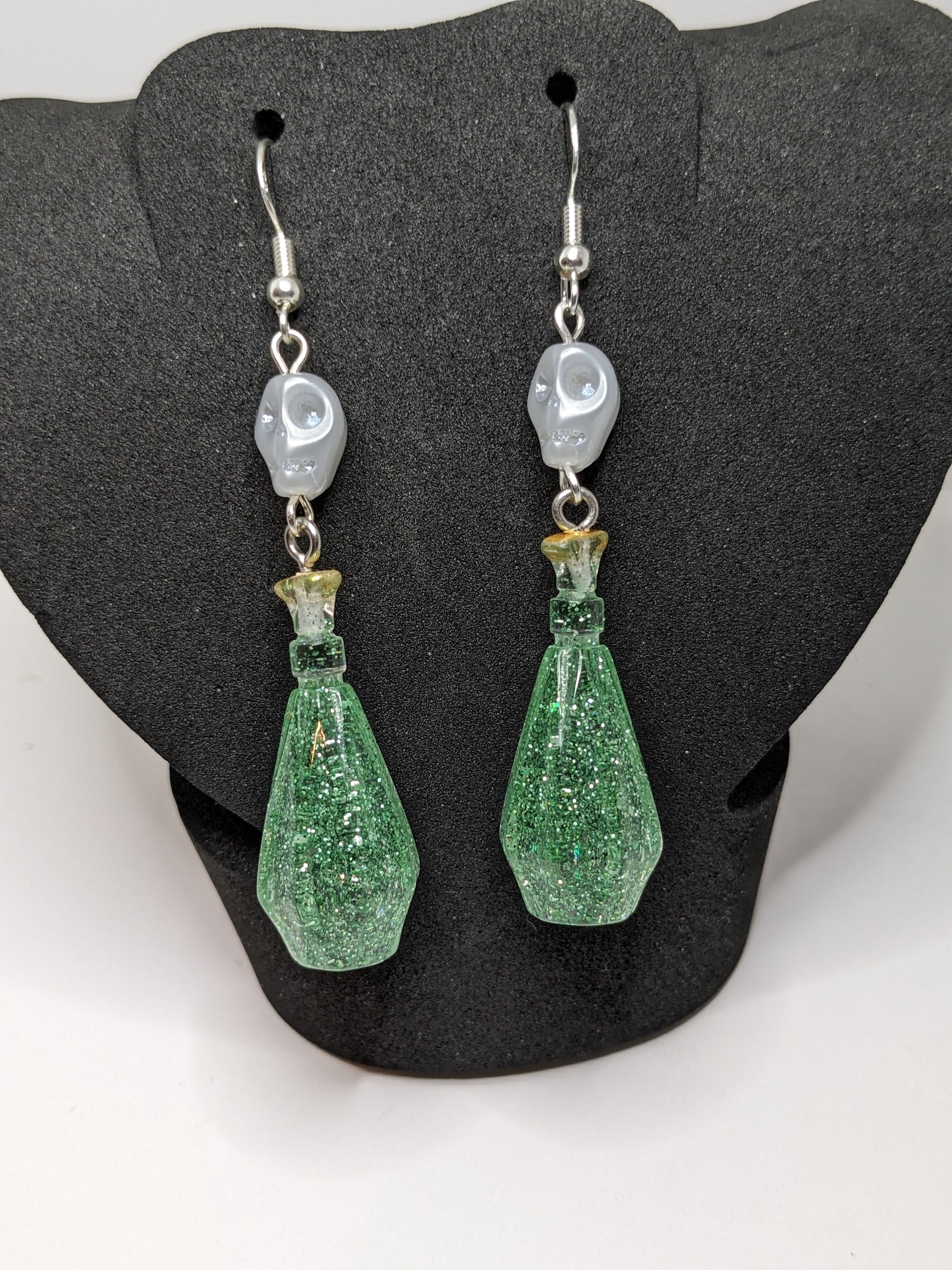 Green glittery potion bottle earrings with white skull beads on a dark gray stand.