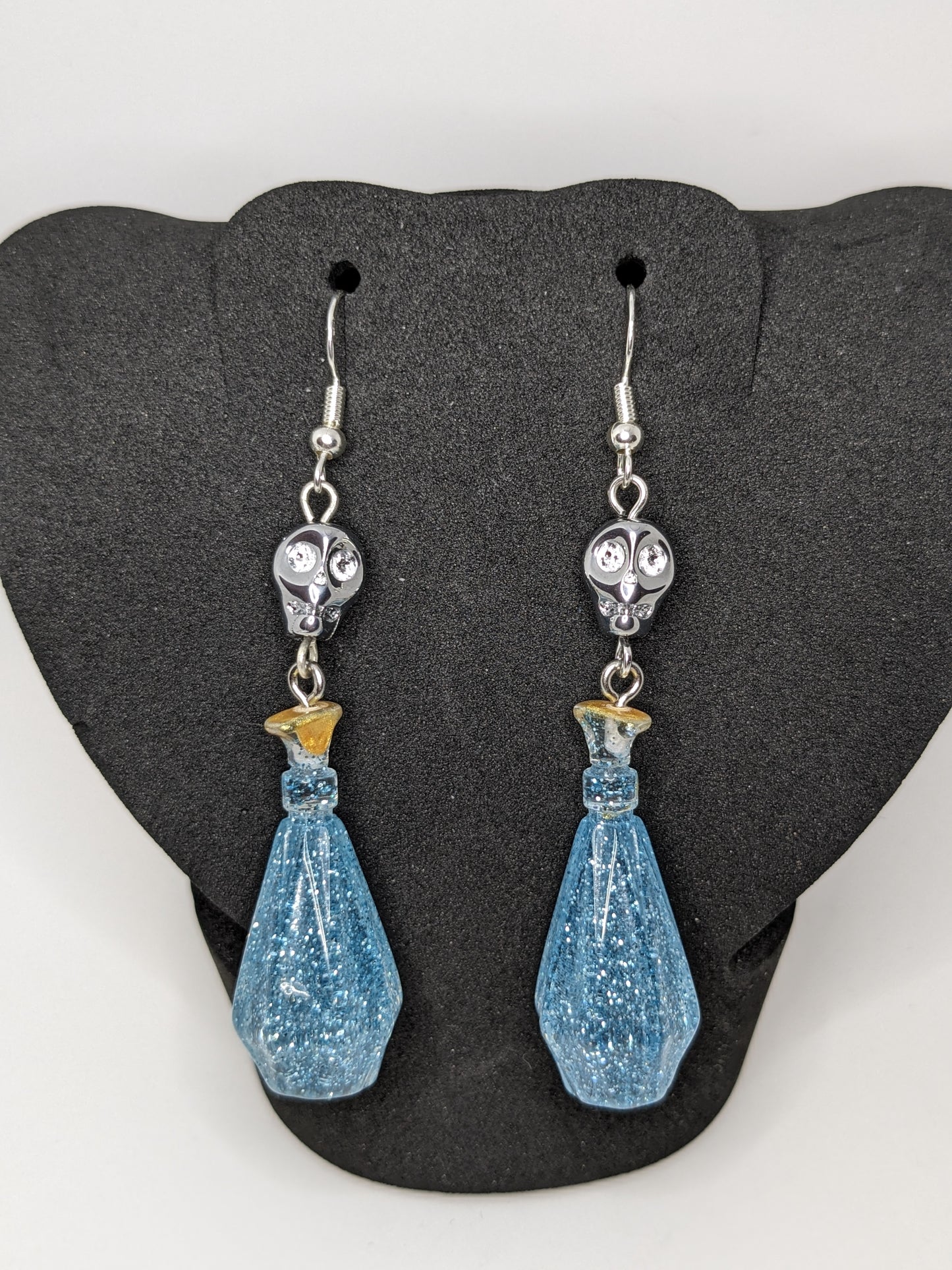 Blue faceted potion bottle shaped earrings with silver skull accents on a black stand.