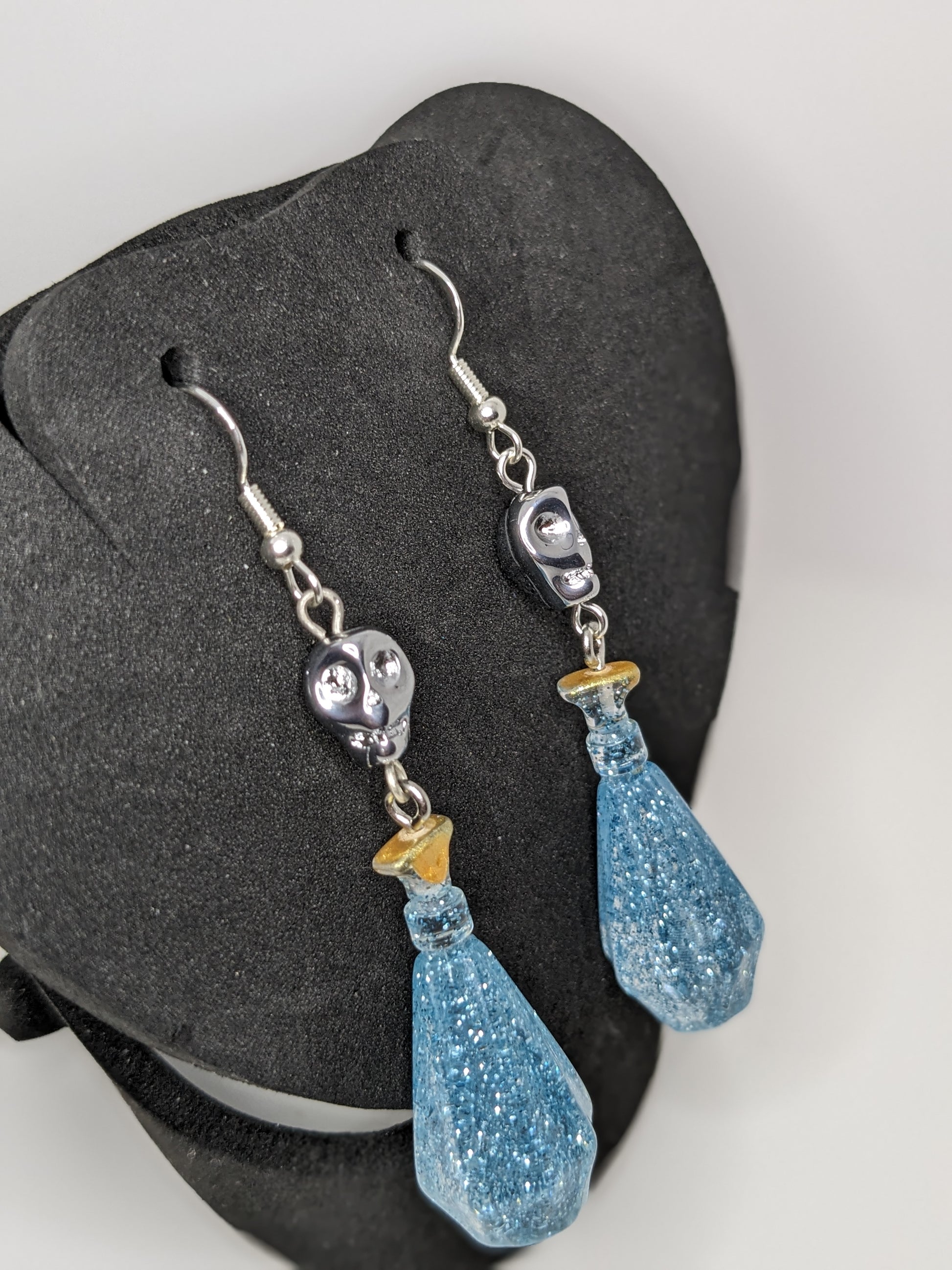 Blue faceted potion bottle shaped earrings with silver skull accents on a black stand.