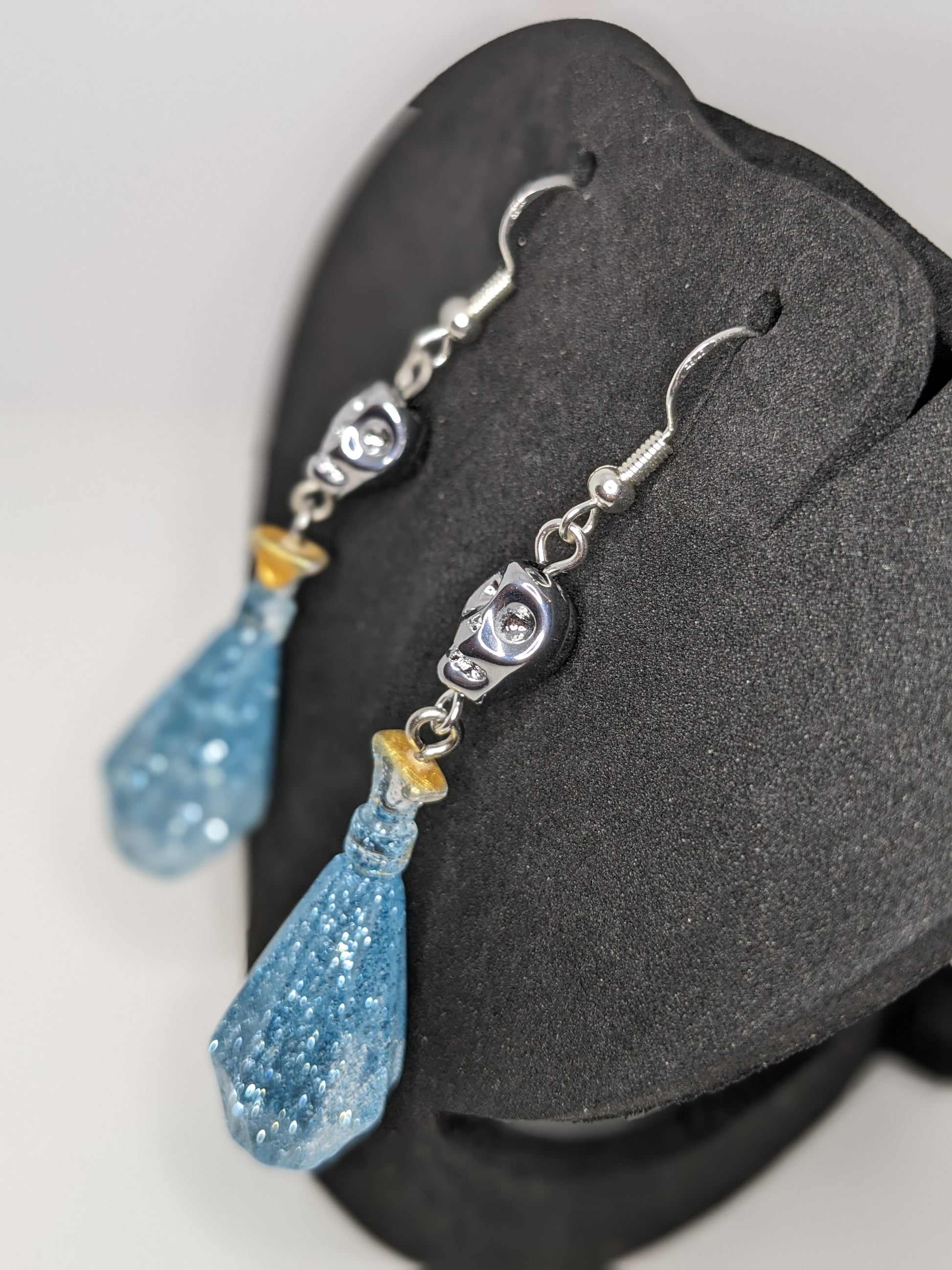 Blue faceted potion bottle shaped earrings with silver skull accents on a black stand.