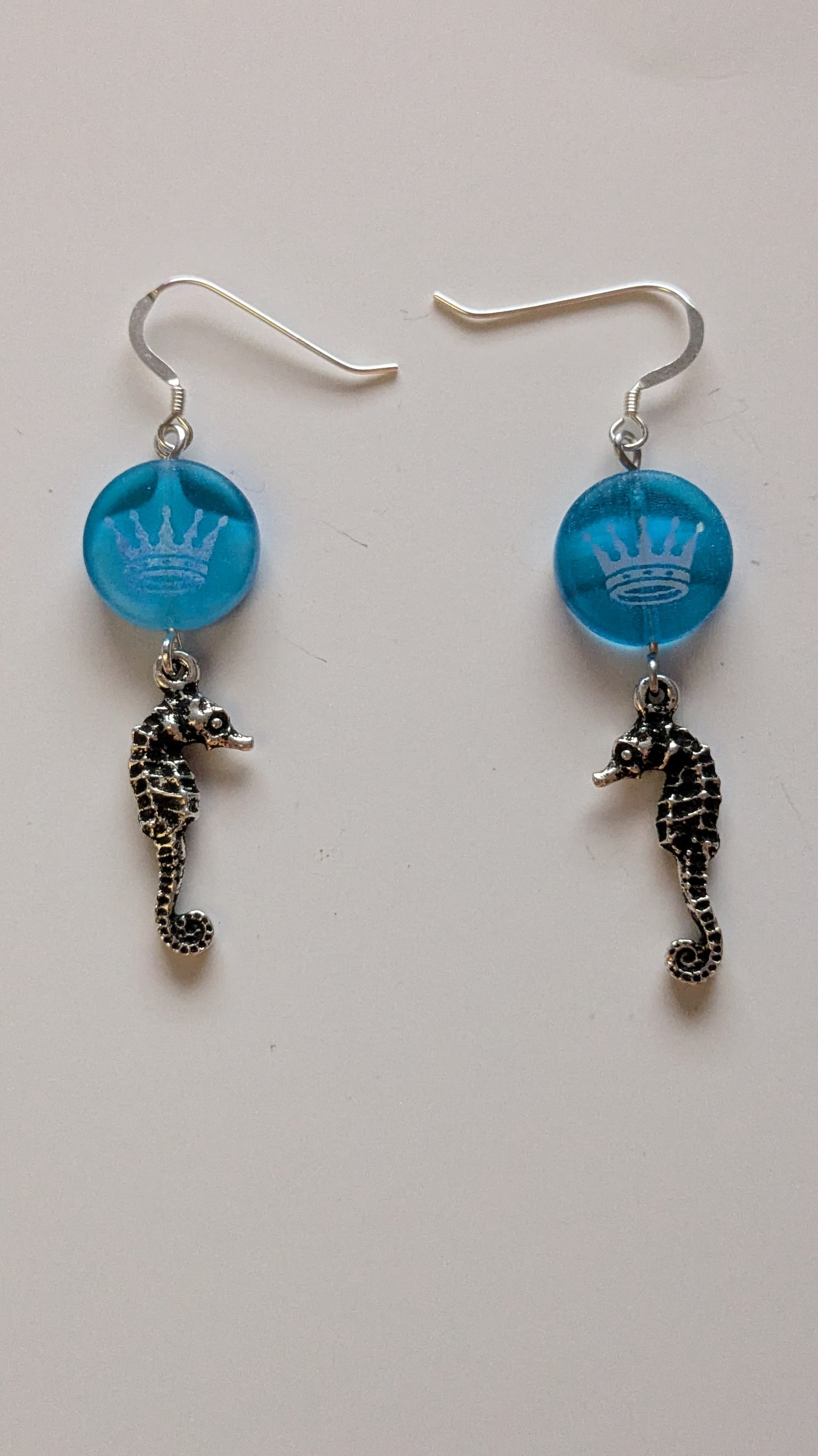 Pair of earrings with blue circular charms with a silver crown overlay and silver seahorse pendants on a light gray background.