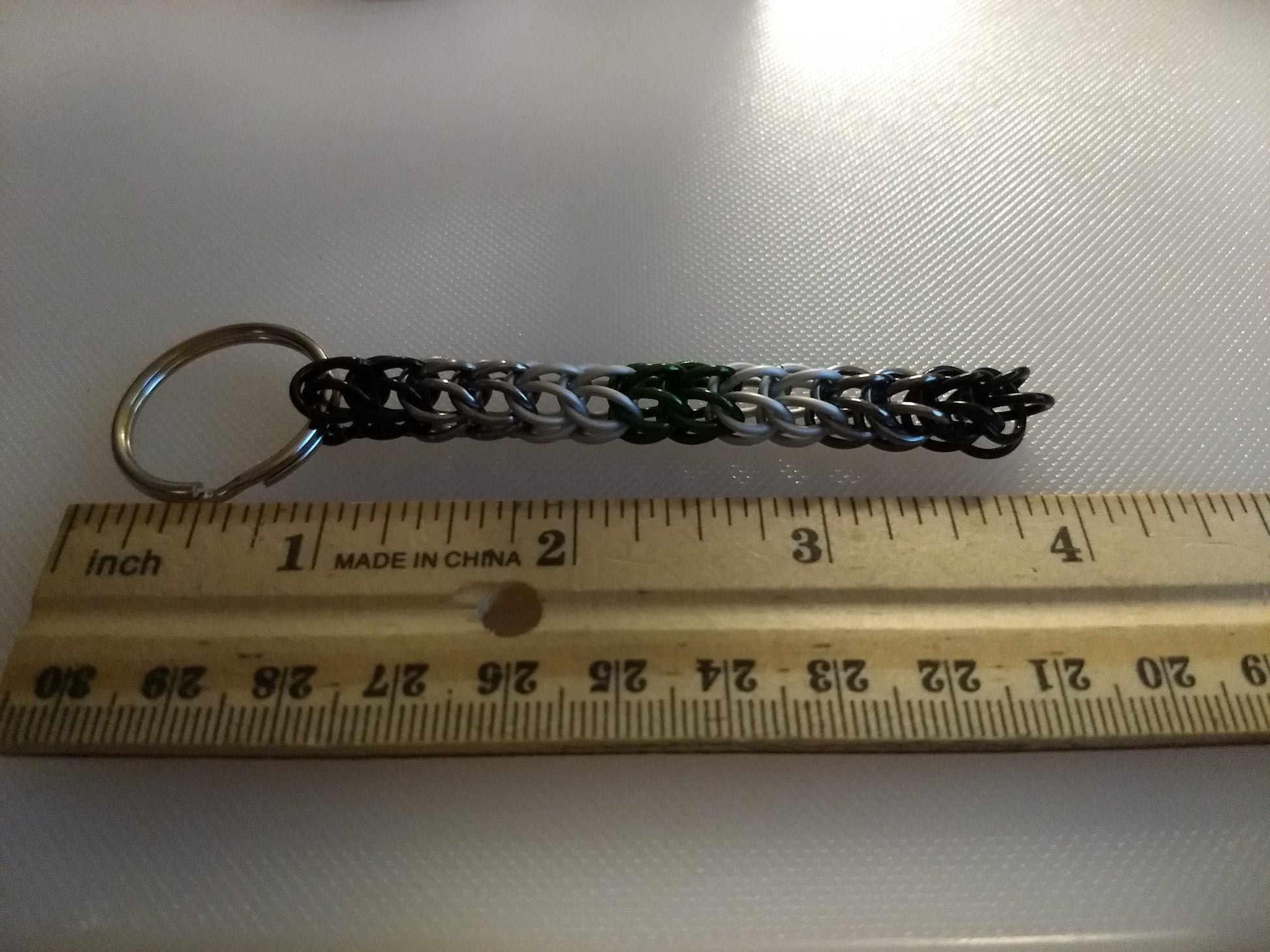 Chainmail keychain in Agender flag colors next to a wooden ruler showing it is 3 inches long
