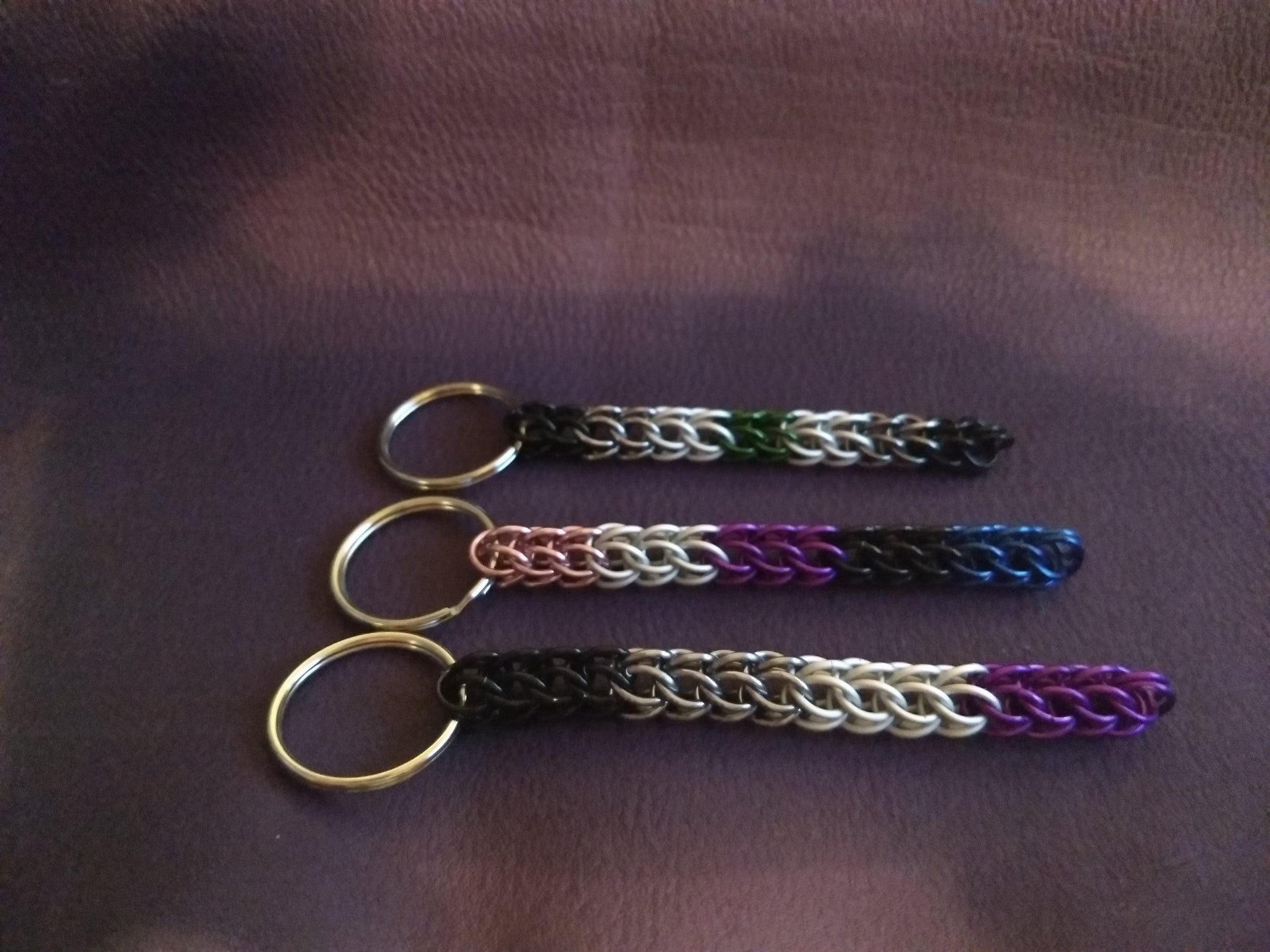 Three chainmail keychains in pride colors on a dark background: Agender, Genderfluid, Asexual