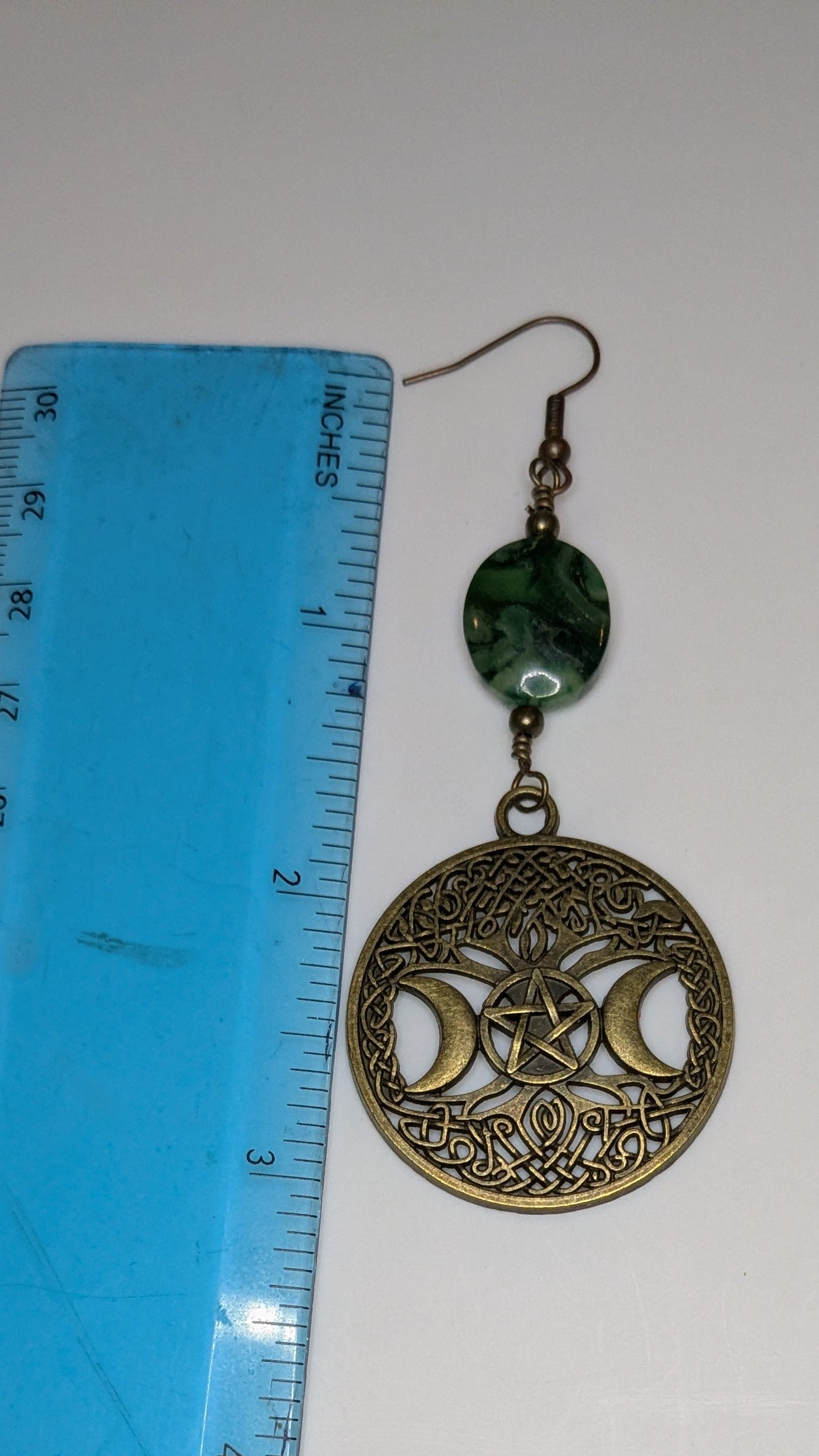 Decorative earring with green bead and blue ruler for scale on a white background