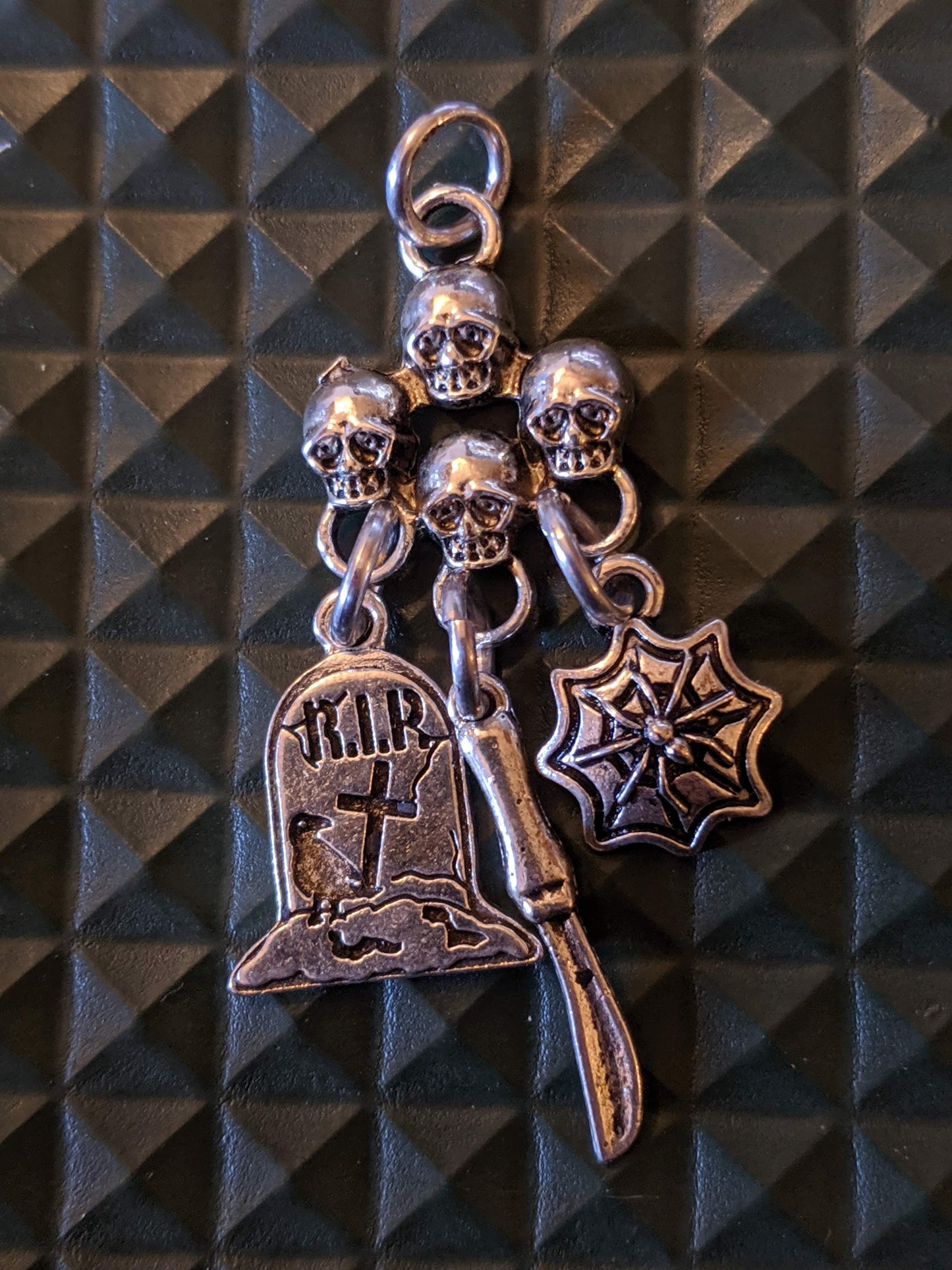 A silver-toned pendant with four skull motifs, featuring charms such as a gravestone, spiderweb, and machete, attached to a black cord.