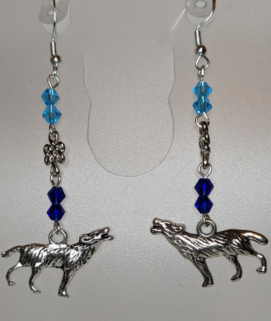 Close up of a pair of earrings with silver howling wolf charms at the bottom and faceted bicone crystal beads in light blue & dark blue separated by a silver Celtic knot connector.