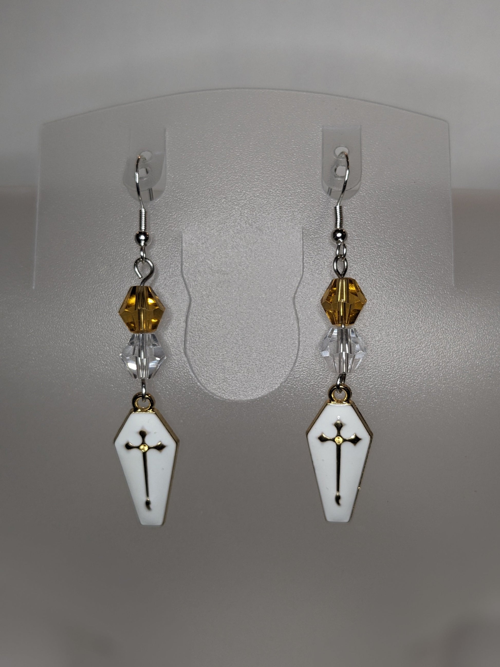 A pair of Vamp Goth Earrings featuring coffin-shaped charms enameled in white with a gold cross with faceted gold and clear bead accents.