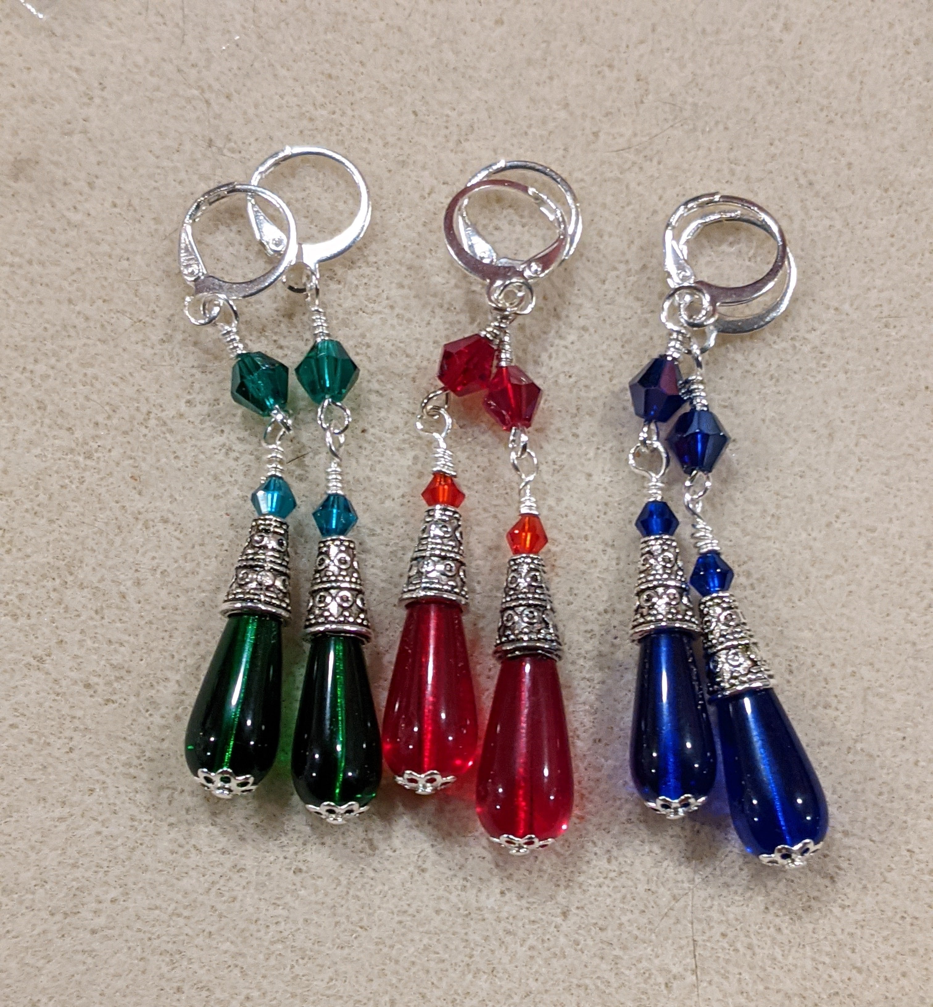 A set of three Czech pressed glass dangle earrings in red, green, and blue, each featuring a teardrop-shaped glass bead and crystal bi cone beads, wire wrapped and dangling from silver ear wires.