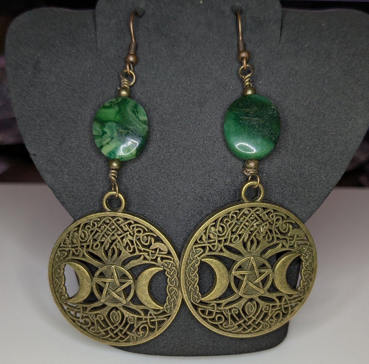 Pair of bronze filigree detailed earrings with green beads on a gray stand.