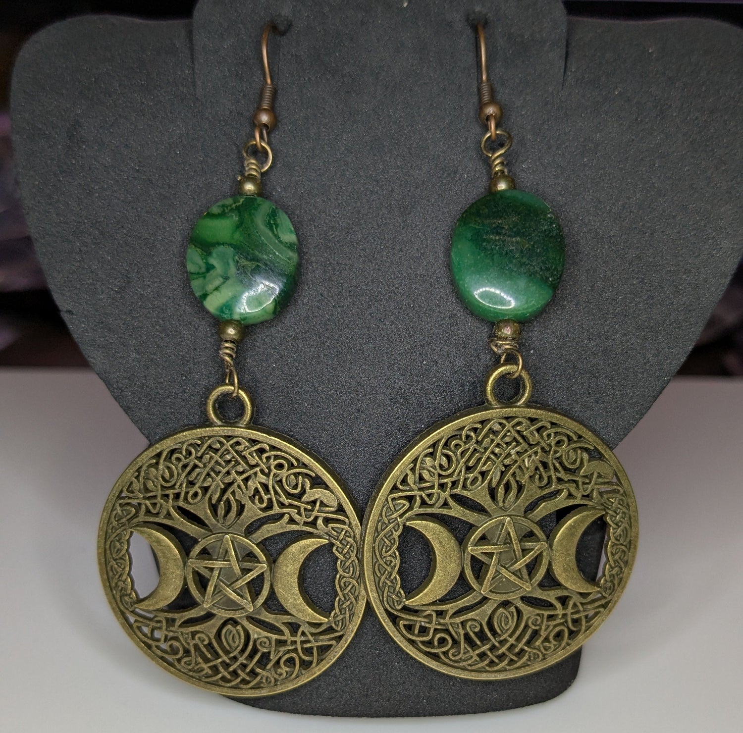 Pair of bronze filigree detailed earrings with green beads on a gray stand.